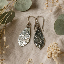Load image into Gallery viewer, Silver earrings on a textured fabric background with dried leaves and flowers.